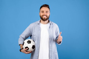 Futbol topu tutan neşeli genç adam, baş parmak hareketi yapıyor, mavi stüdyo arka planında spor öneriyor. Futbol fanatiği portresi şampiyonluk maçında en sevdiği takımı destekliyor.
