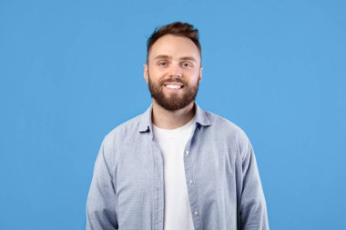 Sıradan giyinmiş, gülümseyen ve mavi arka planda kameraya bakan yakışıklı sakallı genç adamın portresi. Çekici gülüşü olan, mutlu ve arkadaş canlısı bir adam.