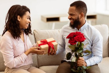 Romantik bir hediye. Siyah adam elinde bir demet çiçek tutuyor ve evinde birlikte oturup tatilini kutlayan karısına paket kutusunu veriyor. Doğum günü Hediyeleri, Tatil Kutlama Konsepti