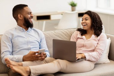 Aile Gadget 'ları. Afro-Amerikan çift koltukta dinleniyor ve birbirlerine bakıyorlar, kadın dizüstü bilgisayar kullanıyor, erkek akıllı telefon tutuyor evde interneti birlikte tarıyor. Modern Yaşam Tarzı Konsepti