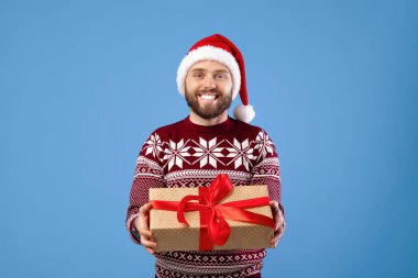 Sıcak süveter ve Noel Baba şapkalı mutlu genç adam mavi stüdyo arka planında hediye kutusu tutuyor. Yılbaşı hediyesi olan olumlu milenyum çocuğu kış tatilini kutluyor.