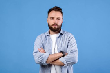 Kollarını kavuşturup mavi stüdyonun arka planındaki kameraya bakan ciddi bir milenyum erkeğinin portresi. Kendine güvenen sakallı, sıradan giyinmiş, havalı bir yüz ifadesi olan genç bir adam.