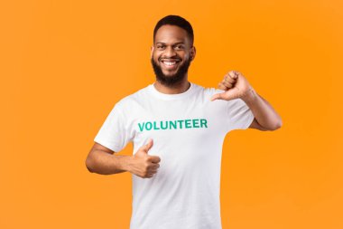 Gönüllü olmayı seviyorum. Gülümseyen Afrikalı gönüllü bir adam kendini işaret ederek sarı stüdyo arka planında poz veriyor, kameraya gülümsüyor. Eylemcilerin portresi