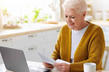 Mutfaktaki modern dizüstü bilgisayarla masada oturan, mektup okuyan, gülen ve alan kopyalayan mutlu bir büyükanne. Elinde belge ya da fatura tutan gülümseyen yaşlı kadın, iyi haberler var, ev içi.