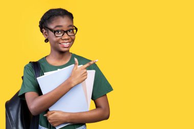 Sarı Stüdyo Arkaplanının üzerinde duran kitap ve defterlerle mesajlaşmak için boş uzayı işaret eden gülümseyen siyah kız öğrenci. Büyük Eğitim Teklif Reklamı