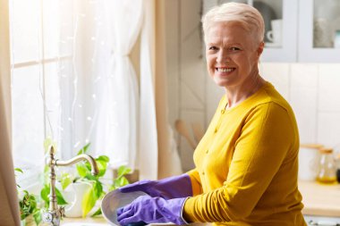 Neşeli, yaşlı, gri saçlı, rahat mutfakta bulaşık yıkayan, ellerini, arka görüşünü, fotokopi alanını korumak için lastik eldiven kullanan bir bayan. Evde bulaşık yıkayan çekici bir büyükanne, bezle tabağı temizliyor.