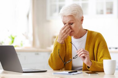 Evde çalışan, dizüstü bilgisayar kullanan, mutfakta oturan, fotokopi çeken, beyaz saçlı, olgun bir kadın. Kıdemli bayan not alıyor, bilgisayarın önünde oturuyor, bardak tutuyor, burun köprüsünü ovuyor.