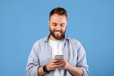 Çevrimiçi yaşam sondası. Mavi stüdyo arka planında cep telefonu kullanan acayip sakallı adam. Modern aletlerle internette gezinen, video izleyen, sosyal medyada paylaşan pozitif genç bir adam.