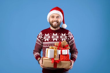 Yünlü süveter ve Noel Baba şapkalı neşeli genç adamın portresi mavi stüdyo arka planında hediyeler barındırıyor. Sakallı milenyum adamı bir yığın hediyeyle yeni yılı kutluyor.