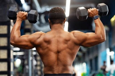 Afrikalı Amerikalı gömleksiz vücut geliştirme uzmanı modern spor salonunda kas egzersizi yapıyor, dambılları kaldırıyor, siyah adam pazıları ve arkası için egzersiz yapıyor.