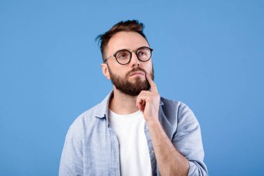 Mavi stüdyo arka planında derin düşüncelere dalmış, gözlüklü, çenesine dokunan dalgın sakallı adamın portresi. Kafası karışmış genç bir adam seçim yapıyor ya da bir şeyler hayal ediyor.