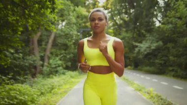 Genç bir Afrikalı Amerikalı kadın yeşil bir parkta koşuyor ve spor rutinini aktif olarak uyguluyor. Parlak atletik kıyafetler giyiyor, açık havadan ve sağlıklı bir yaşam tarzından hoşlanıyor..