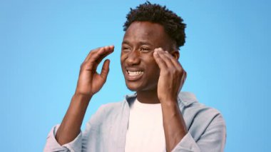 Genç bir Afro-Amerikan adam kameraya bakmıyor ve kafasına dokunuyor. Beyaz bir tişörtün üzerine açık renk bir tişört giyer, parlak mavi bir arka plana yaslanır, boşluğu taklit eder.
