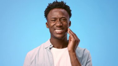 Mutsuz genç Afro-Amerikan bir adam parlak mavi bir fon karşısında duruyor, diş ağrısı çekiyor, eliyle yanağına dokunuyor.