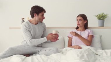 Genç bir çift yatakta birlikte oturur, sohbet ederler. Kadın endişelenirken, erkek elinde bir bardak tutuyor, vücut ısısını kontrol ediyor.