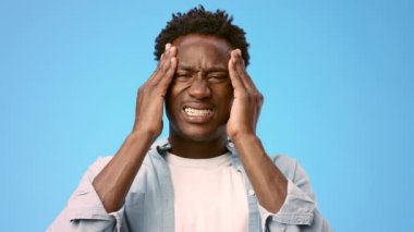 Afrika kökenli genç bir adam, parlak mavi bir arkaplana karşı başını tutarken hayal kırıklığı sergiliyor. Yüzünde şiddetli bir baş ağrısı ya da migren var.