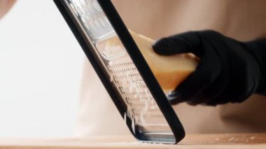 Minimalist bir mutfakta yetenekli bir şef siyah eldiven giyerken bir kalıp peyniri titizlikle rendeliyor. Odaklanmış atmosfer, sağlıklı yemek pişirme ve sürdürülebilirliğe olan bağlılığı vurguluyor.