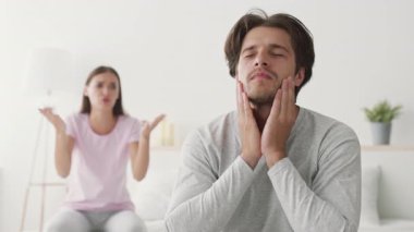 Bir erkek, elleri yüzü üzerinde görünür bir şekilde stresli bir şekilde yatakta otururken, bir kadın arka planda elleri ile hareket eder. Parlak dekor o anın duygusal atmosferini arttırır..