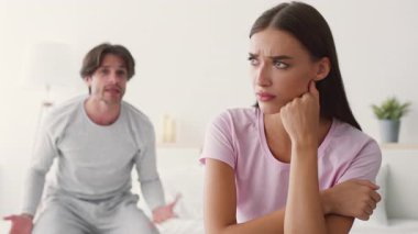 Çağdaş bir yatak odasında, bir kadın üzgün ve mesafeli görünürken bir erkek hayal kırıklığını ifade eder. Gerginlik, çift arasında kişisel alanlarında çözülmemiş sorunlara işaret ediyor..