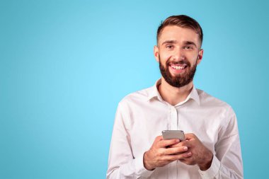 Cheerful millennial guy with smartphone browsing web, playing game, shopping online on blue studio background. Positive Caucasian man with modern gadget looking at camera and smiling
