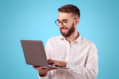 Resmi giyinen başarılı genç adam mavi stüdyo arka planında dizüstü bilgisayar kullanıyor. Çevrimiçi işi olan neşeli iş adamı, internet konferansına katılıyor, internet üzerinden iletişim kuruyor.