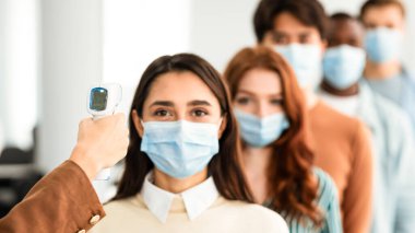 Sıraya dizilmiş çeşitli insanlar için ateşi ölçen sıcaklığı ölçen kadın, kapalı alanda kızıl ötesi temassız Themometer kullanıyor. Coronavirus, Covid-19 Salgın Konsepti
