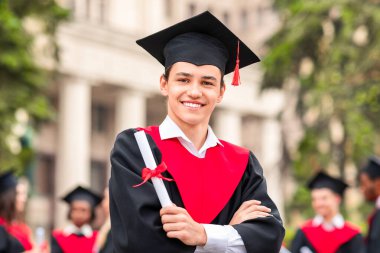 Mezuniyet kostümlü neşeli adam diplomasını gösteriyor ve kameraya gülümsüyor. Üniversite kampüsünde uluslararası öğrenci grubuna poz veren mutlu bir erkek öğrenci.