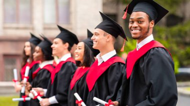 Mezuniyet törenine katılan çeşitli uluslararası öğrenciler mezuniyet şapkası ve cüppeli mutlu çok ırklı öğrenciler üniversite kampüsünde sıraya dizilmiş.