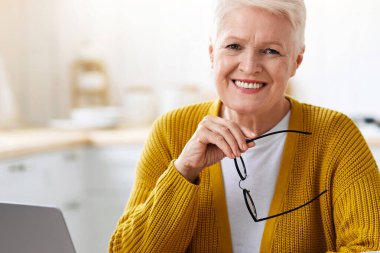 Çevrimiçi eğitim, kurslar, internet konsepti. Mutfakta dizüstü bilgisayarın önünde oturan, elinde bardaklar olan, gülümseyen, çay içen, internetten ders çalışmaktan keyif alan mutlu son sınıf kadını.