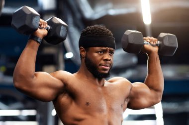 Kendine güvenen Afro-Amerikan tişörtsüz vücut geliştirici spor salonunda kas egzersizi yapan, dambılları kaldıran, pazılarını ve sırtını çalıştıran, fotokopi alanına bakan...