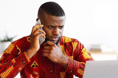 Kabile gömlekli meşgul bir iş adamının portresi cep telefonuyla konuşuyor ve dizüstü bilgisayarla çalışıyor, Afrikalı girişimci geleneksel kıyafetlerle bilgisayar ekranına bakıyor ve telefon görüşmesi yapıyor.