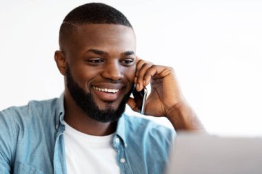 Cep telefonuyla konuşan ve ofiste dizüstü bilgisayar kullanan gülümseyen siyah adamın yakın plan portresi. Yakışıklı Afrikalı Amerikalı Girişimci Bilgisayar ve Seçici Odaklanma Üzerinde Çalışırken Telefon Konuşmasından Hoşlanıyor