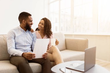 Aile Bütçesi Planlama. Gülümseyen Afro-Amerikan adam elinde kağıt, çalışıyor ve evde belgeler okuyor, kadın kahve içiyor ve ona bakıyor, dizüstü bilgisayarla kanepede oturuyor.