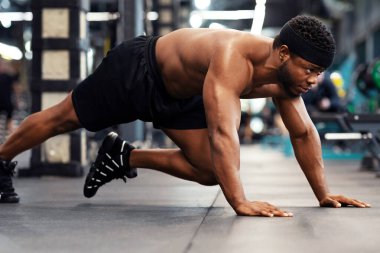Plank konsepti işe yarıyor. Modern erkek spor salonunda spor yapan etkin ve üstsüz bir zenci. Motive olmuş Afro-Amerikan kaslı vücut geliştirici dizlerinin üstüne çökmüş, panorama