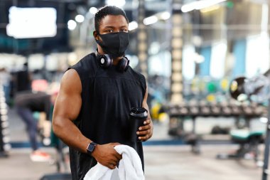 Spor ve karantina. Koruyucu maskeli Afrikalı Amerikalı vücut geliştirici spor salonunda su ya da protein ve havlu tutuyor, boş alan. Modern spor salonunda egzersiz yaparken siyah kaslı adam mola veriyor.