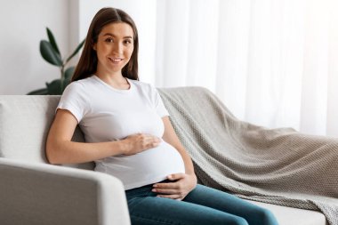 Evdeki kanepede oturan hamile genç bayan karnını kucaklıyor ve kameraya gülümsüyor. Güzel, güzel bir kadının oturma odasında dinlenmesini ve hamileliğin tadını çıkarmasını bekliyorum.