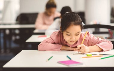 Okul harcı konsepti. Sınıfta oturan, not defterinde yazan ya da çizen, ödev yapan ya da ders sırasında not alan, seçici odak, ücretsiz kopya alanı olan küçük Asyalı kız portresi