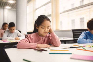 İlkokulda ya da anaokulunda sınıftaki masada oturan, defterde okuma ödevi yapan odaklanmış küçük Asyalı kız portresi. Coronavirus karantinasından sonra tekrar açılıp okula geri dönmek.