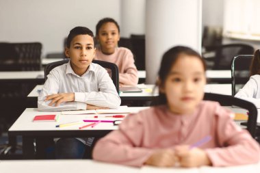 Okulun ilk günü. Odaklanmış, çalışkan bir çocuğun portresi. Kollarını kavuşturmuş, arka planda bulanık sınıf arkadaşlarıyla oturuyor. Öğretmeni dinliyor, seçici odaklanıyor. Çalışmaya Geri Dön