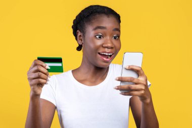 Mobil Alışveriş. Heyecanlı Siyah Kadın Kredi Kartı ve Akıllı Telefon Tutuyor, Dükkanlar Çevrimiçi Başvuru Vatandaşları Sarı Stüdyo Arkaplanının Üzerinde Durmakta. Heyecanlı Müşteri, Alışveriş Uygulaması ve Nakit