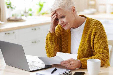 Faturaları kontrol ederken kafasını tutan yaşlı bayanın karantina sırasında maddi sorunları vardı. Mutfakta dizüstü bilgisayarın önünde oturan üzgün yaşlı kadın ve elinde belgeler, boşluğu kopyala