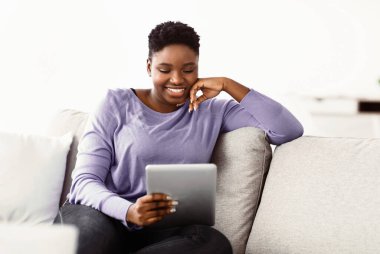 Dinlen ve rahatla kavramı. Neşeli kıvrımlı zenci kadın film izliyor, cep telefonuyla kayıt yapıyor. Rahat Afro-Amerikan genç bayan kanepede oturuyor, dijital tablet kullanıyor,