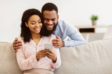 Gülümseyen genç siyah adam akıllı telefona bakarken kadını arkadan kucaklıyor. Afrika kökenli Amerikalı çift sosyal medyayı cep telefonuyla paylaşıyor, mutlu kadın kanepede oturuyor, aletleri gösteriyor.