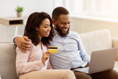 Çevrimiçi Alışveriş, Ticaret Konsepti. Mutlu Afrikalı genç Amerikalı çift dizüstü bilgisayar kullanıyor ve internetten kıyafet veya ürün alıyor, bankamatik kartıyla ödeme yapıyor, evdeki kanepede oturuyor.