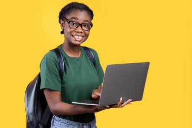 Mutlu Siyahi Öğrenci Kız Laptop Bilgisayar Kullanıyor Kameraya Gülümsüyor Sarı Arkaplanda Duruyor. Stüdyo çekimi. E-Öğrenme ve Çevrimiçi Eğitim. Eğitim Aletleri Konsepti