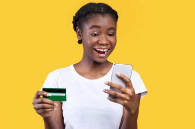 Alışverişe. Neşeli Afrikalı Amerikalı Kadın Telefon Tutma Kredi Kartı Kullanıyor, Dükkanlar Çevrimiçi Cep Telefonu Uygulaması Sarı Arkaplanda Duruyor. Stüdyo çekimi. Alışveriş Uygulaması Konsepti