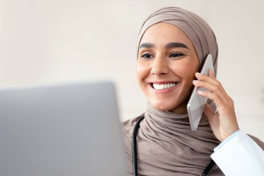 Gülümseyen kadın doktorun telefon görüşmesi yapması, dizüstü bilgisayar kullanması, klinik içini kullanması, fotokopi çekmesi. Başörtülü güzel Müslüman kadın hasta ya da meslektaşıyla cep telefonundan konuşuyor.