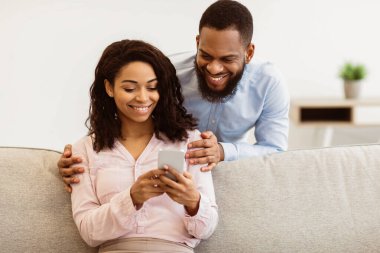 Neşeli milenyum siyahi çifti akıllı telefondan internete girip birlikte video izliyorlar. Yakışıklı erkek arkadaşı arkadan kız arkadaşını kucaklıyor, kadın kanepede oturuyor.