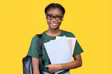 Sarı Stüdyo arka planında duran ve kameraya gülümseyen kitaplı mutlu siyah kız öğrenci pozu. Üniversite ve Üniversite Eğitimi, Öğrencilerin Yaşam Tarzı Konsepti