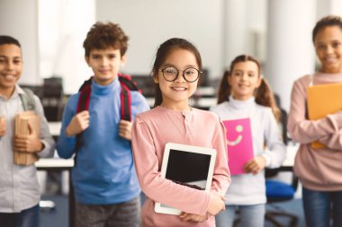 Öğrenciler, Teknoloji ve Öğrenme Konsepti. Gözlüklü gülümseyen Asyalı kız elinde dijital tabletle arka planda bir grup çok kültürlü sınıf arkadaşıyla duruyor ve ilkokulda okuyor.